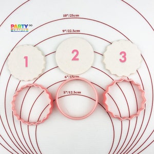 Op de afbeelding: Drie witte fondantcirkels met de nummers 1, 2 en 3 erop gedrukt. Drie roze koekjesuitstekers met gegolfde randen, met een diameter van 12,5 cm, 15 cm en 22,5 cm. De koekjesuitstekers en fondantcirkels bevinden zich op een wit oppervlak met concentrische rode cirkels.