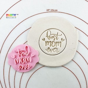 Puede incluir: Cortador de galletas rosa y blanco con el texto "Best Mom Ever" en una fuente cursiva. El cortador está sobre una superficie blanca con un círculo marrón alrededor.