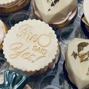 Puede incluir: Primer plano de un cupcake blanco con un adorno de azúcar decorativo que dice "She Said Yes!" en letras doradas. El cupcake está rodeado de otros cupcakes con diferentes decoraciones.