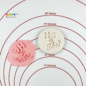 Puede incluir: Dos cortadores de galletas de silicona rosa y blanco con el texto "He or She?" en ellos. Los cortadores están sobre una superficie blanca con círculos concéntricos rojos.