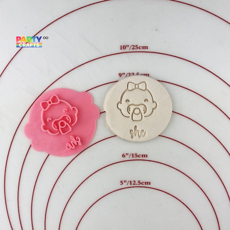 May include: Two pink and white silicone baby cookie cutters with the word "she" on each. The cutters are on a white surface with red concentric circles.