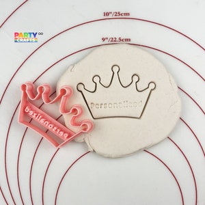 Pode incluir: Cortador de biscoitos de plástico rosa em forma de coroa com a palavra "Personalizado" impressa no cortador. O cortador está sobre uma superfície branca com círculos concêntricos vermelhos.