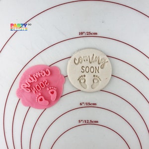 May include: Two pink and white silicone baby shower cookie cutters. The larger cutter is oval shaped and reads "coming soon" with two small footprints. The smaller cutter is shaped like a cloud and reads "primo 2002" with two small footprints.