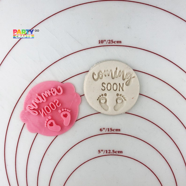 May include: Two pink and white silicone baby shower cookie cutters. The larger cutter is oval shaped and reads "coming soon" with two small footprints. The smaller cutter is shaped like a cloud and reads "primo 2002" with two small footprints.