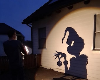 May include: A projection light device casts a large, dark shadow of a character with a tall, curved head and long fingers holding a round ornament onto the white exterior wall of a house during the evening hours.