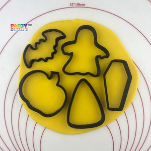 Pode incluir: Uma folha de fondant amarela com cinco cortadores de biscoitos de Halloween: um fantasma, um morcego, um caixão, um milho doce e uma abóbora. Os cortadores são feitos de plástico preto.
