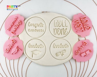 May include: A set of six pink and white cookie cutters with graduation themes. The cutters feature the phrases "Congrats Graduate", "Well Done", "Graduate", and "Class of 2023".