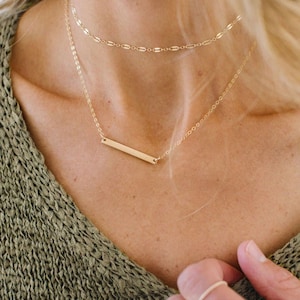 May include: A gold bar necklace with a delicate chain. The necklace is layered with a second, thinner chain.