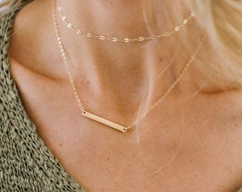 May include: A gold bar necklace with a delicate chain. The necklace is layered with a second, thinner chain.