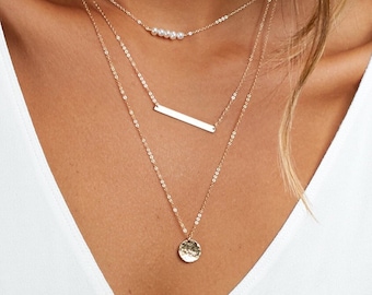 Puede incluir: Un collar de oro de varias capas con un pequeño colgante de barra y un colgante circular martillado. La capa superior presenta una fila de pequeñas perlas blancas.