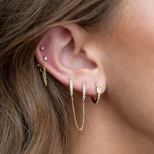 May include: An ear featuring several gold earrings. There are small stud earrings, chain earrings, and hoop earrings with clear gemstones. The jewellery is set against the backdrop of the ear and hair.