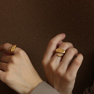 May include: Two gold rings, one with a curved oval shape and the other with a straight oval shape, are worn on the fingers of a person's hand.