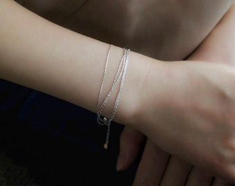 May include: A delicate silver-toned multi-strand bracelet featuring thin, fine chains that wrap around the wrist. The bracelet includes a small, dangling charm at the end of the adjustable chain, providing a layered appearance on the wrist.