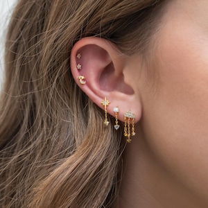 May include: Gold earrings with a cloud charm and dangling stars. The earrings are worn in a person's ear with other earrings, including a crescent moon and a star.