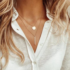 May include: A gold layered necklace with a round pendant and a long, thin bar pendant.