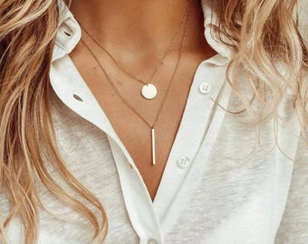 May include: A gold layered necklace with a round pendant and a long, thin bar pendant.