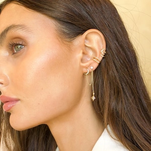 May include: An ear is adorned with a selection of gold-coloured earrings. The set includes a crescent moon stud, a dangling chain with a star charm, a small square stud, and a double-banded ear cuff. The earrings are embellished with small, sparkling stones.
