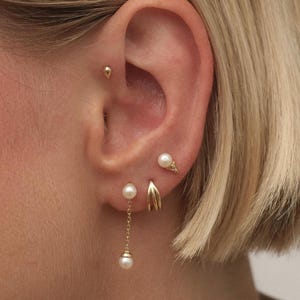 May include: An ear featuring several gold earrings. There's a small stud, a pearl stud, a gold hoop, a pearl drop earring, and a cone-shaped earring with a pearl. The ear is positioned against a backdrop of blonde hair, creating a contrast.