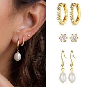 May include: A set of gold earrings with pearl and crystal accents. The set includes a pair of hoop earrings with crystals, a pair of stud earrings with crystals, and a pair of drop earrings with pearls.