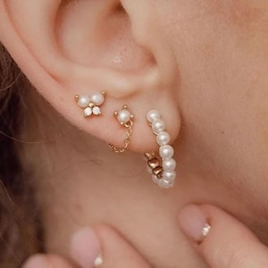 May include: A set of three gold-toned earrings worn in the earlobe. The first is a butterfly-shaped stud with white pearls and clear stones. The second is a pearl stud connected by a gold chain to a hoop. The third is a hoop earring with white pearls.