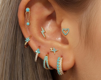 May include: An assortment of gold-coloured earrings featuring turquoise and crystal embellishments. The earrings include a chain drop, a lightning bolt, a heart, a star, and floral designs. Several hoop earrings are also visible, showcasing the variety of styles.