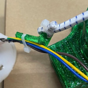 May include: Green 3D printed cable organizer with four wires running through it. The wires are brown, yellow, blue, and green. The organizer is attached to a white bungee cord.