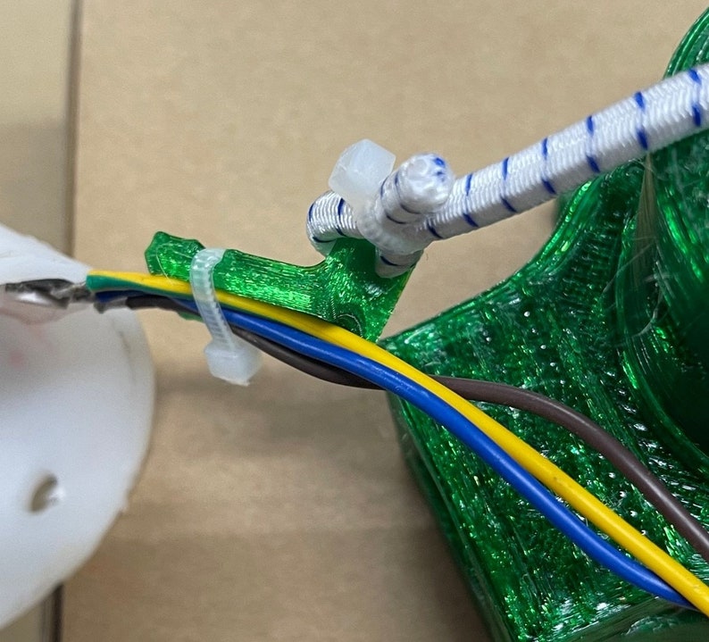May include: Green 3D printed cable organizer with four wires running through it. The wires are brown, yellow, blue, and green. The organizer is attached to a white bungee cord.