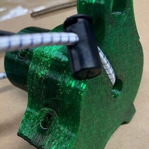 May include: A close-up of a green 3D printed object with a black plastic cap. The object has a hole in the side and a white cord is threaded through the hole.