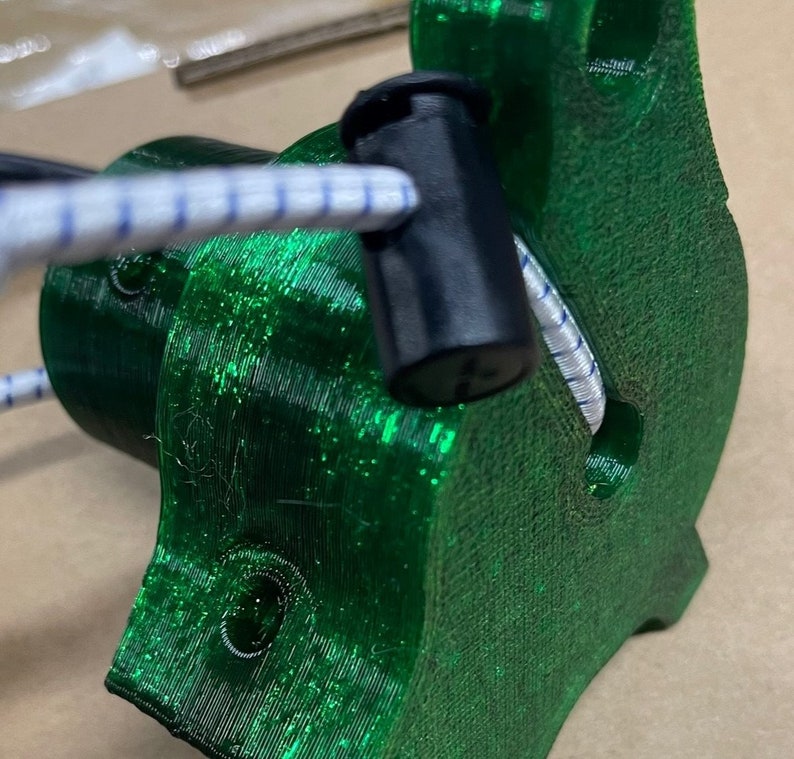 May include: A close-up of a green 3D printed object with a black plastic cap. The object has a hole in the side and a white cord is threaded through the hole.
