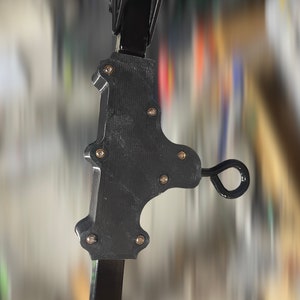 May include: A black, mechanical component with a textured surface, secured with multiple silver-colored fasteners. The component has a curved shape with a loop at the end, and is attached to a black, rectangular support structure.