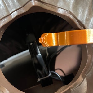 May include: Close-up of the inside of a brown plastic container with a black metal bracket and wires. An orange plastic piece is sticking out of the container.