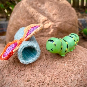 Puede incluir: Un gorro de orejas de conejo de ganchillo azul claro con detalles en naranja, rosa y morado, junto a una figura de oruga verde con marcas negras y amarillas. Los artículos se exhiben sobre una superficie de piedra de color marrón rojizo.