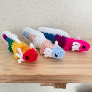 May include: Three crocheted axolotl plushies in different colour combinations. The first is rainbow, the second is pink, blue, and white, and the third is pink and purple.