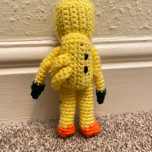 May include: A crocheted yellow plush toy with black eyes and a green and orange design. The toy is standing on a carpet.
