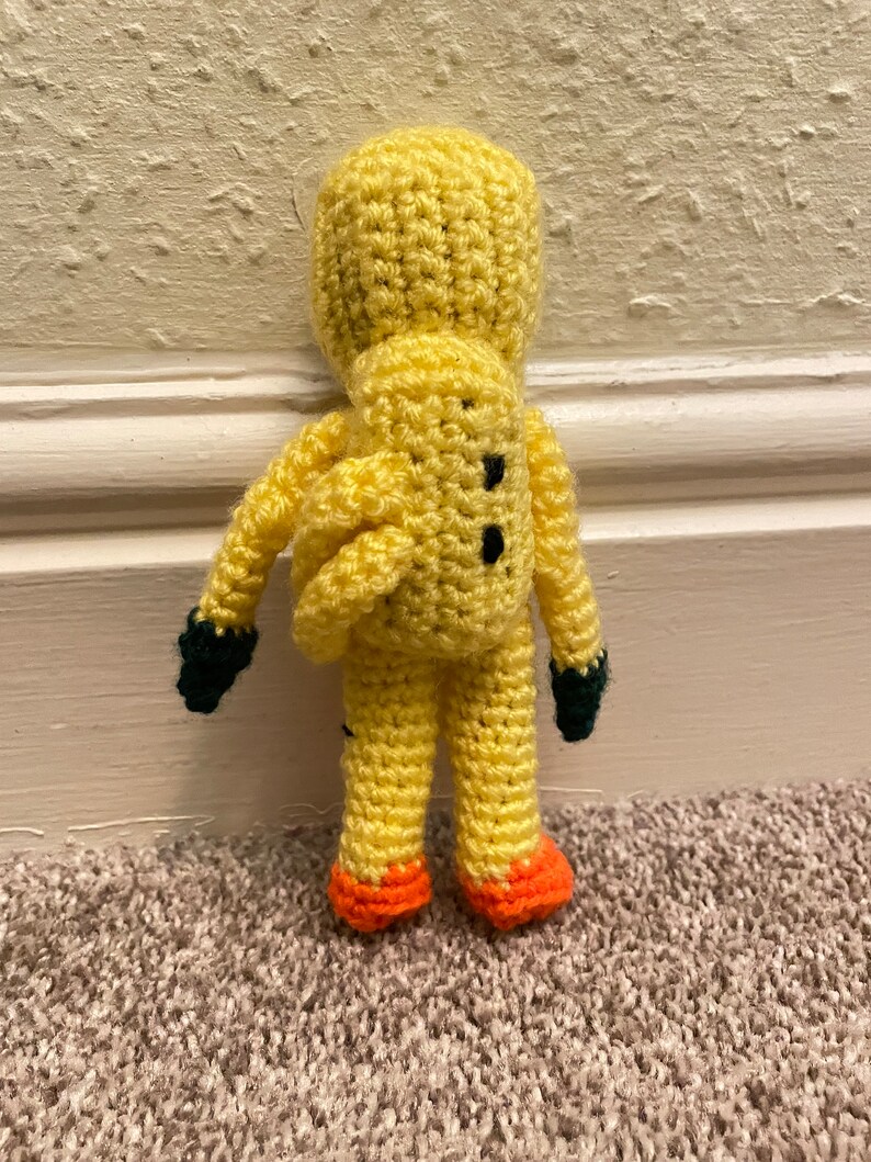 May include: A crocheted yellow plush toy with black eyes and a green and orange design. The toy is standing on a carpet.