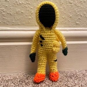 May include: A crocheted yellow doll with a black face mask, black arms, and orange feet. The doll is wearing a yellow hooded jumpsuit.