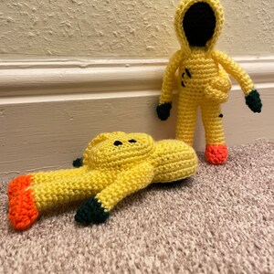 May include: Two crocheted yellow figures wearing hazmat suits. One figure is standing upright, while the other is lying on its back. Both figures have black eyes and orange feet.