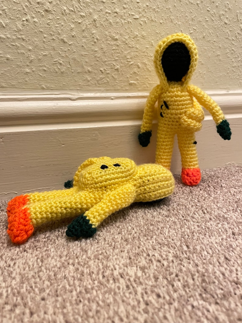 May include: Two crocheted yellow figures wearing hazmat suits. One figure is standing upright, while the other is lying on its back. Both figures have black eyes and orange feet.