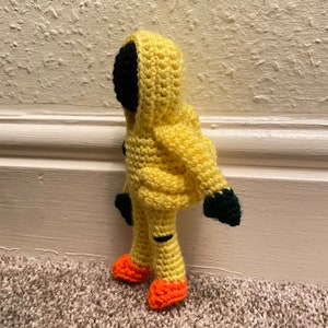 May include: A crocheted yellow hooded figure with black eyes and orange shoes. The figure is standing on a brown carpet.