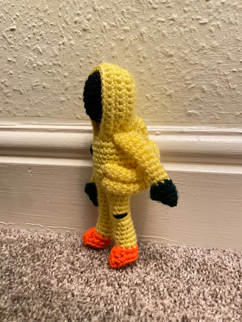 May include: A crocheted yellow hooded figure with black eyes and orange shoes. The figure is standing on a brown carpet.
