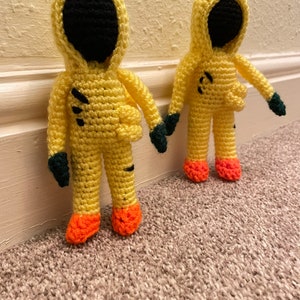May include: Two crocheted yellow figures with black faces and green gloves. The figures are wearing yellow hooded jumpsuits with orange feet.