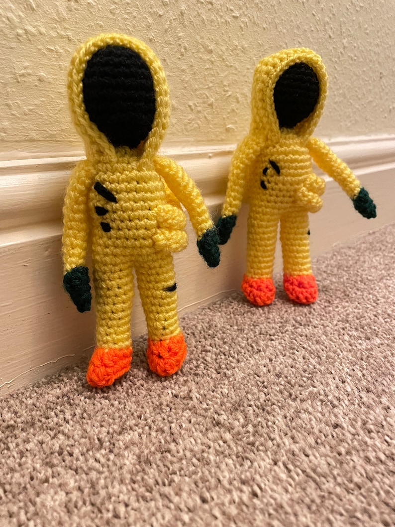 May include: Two crocheted yellow figures with black faces and green gloves. The figures are wearing yellow hooded jumpsuits with orange feet.
