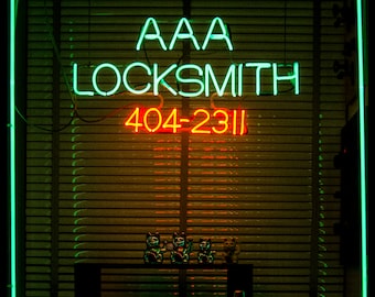 May include: A neon green sign with the text "AAA LOCKSMITH 404-2311" in red neon letters. The sign is in a window with blinds behind it.