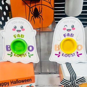 Op de afbeelding: Twee witte spookvormige Halloween-decoraties met de tekst "You are FAB BOO LOUS!". E&eacute;n heeft een groene cirkel, de andere een gele cirkel. De decoraties staan op oranje en witte dozen.