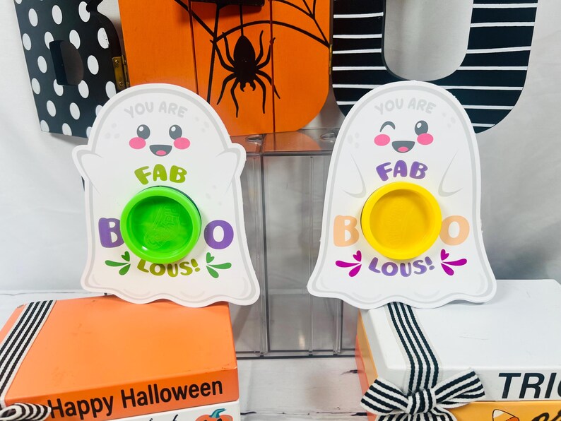 Op de afbeelding: Twee witte spookvormige Halloween-decoraties met de tekst "You are FAB BOO LOUS!". E&eacute;n heeft een groene cirkel, de andere een gele cirkel. De decoraties staan op oranje en witte dozen.