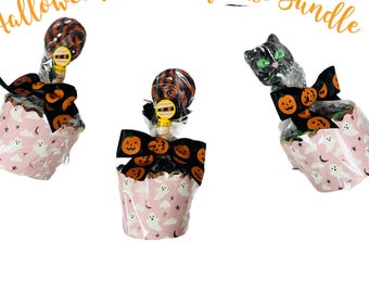 Puede incluir: Tres paquetes de cupcakes de calcetines con temática de Halloween. Cada cupcake está envuelto en papel rosa con diseños de fantasmas y estrellas. Están decorados con un lazo negro con diseños de calabazas naranjas, una piruleta y una figura de gato negro.