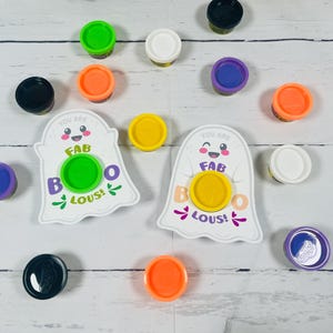 Op de afbeelding: Een verzameling kleurrijke Play-Doh-containers en twee witte spookvormige kaarten met de tekst "You are FAB BOO LOUS!". De Play-Doh-deksels zijn in verschillende kleuren, waaronder groen, oranje, paars, geel, zwart en wit.