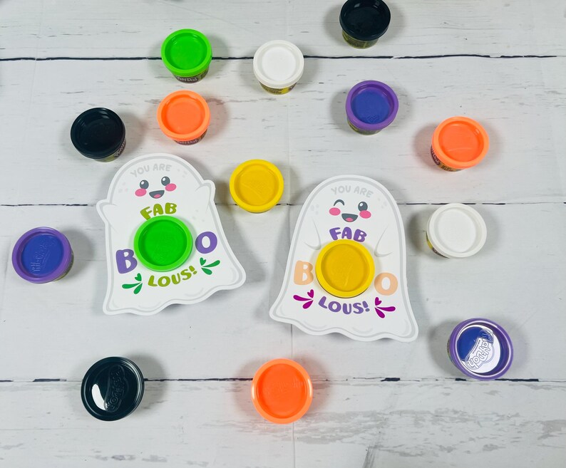 Op de afbeelding: Een verzameling kleurrijke Play-Doh-containers en twee witte spookvormige kaarten met de tekst "You are FAB BOO LOUS!". De Play-Doh-deksels zijn in verschillende kleuren, waaronder groen, oranje, paars, geel, zwart en wit.