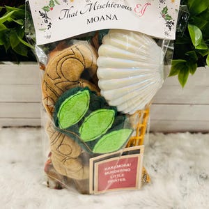 May include: A clear plastic bag filled with various items, including a brown felt toy shaped like a seashell, a green felt toy shaped like leaves, a white seashell, and a red and white sticker that says "Kakamora! Murdering Little Pirates". The bag has a label that says "That Mischievous Elf Moana".