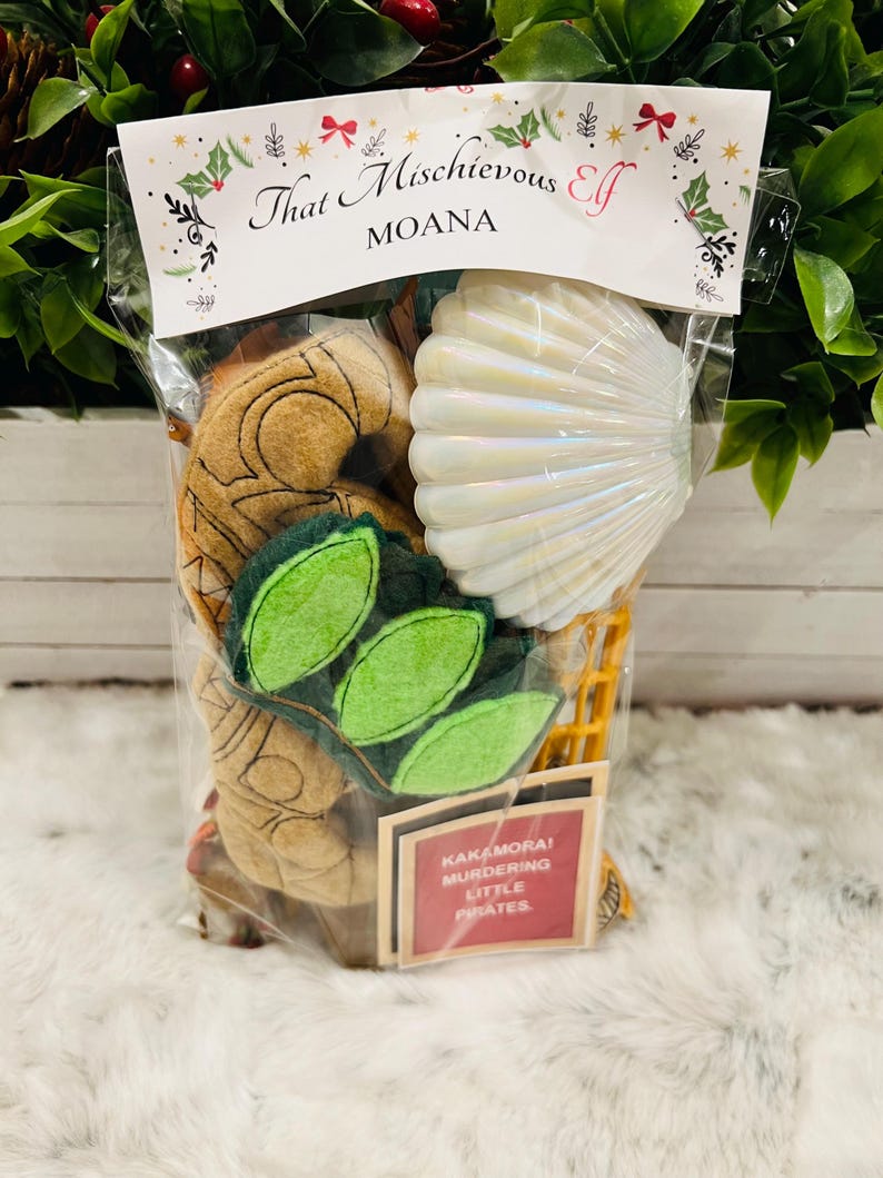 May include: A clear plastic bag filled with various items, including a brown felt toy shaped like a seashell, a green felt toy shaped like leaves, a white seashell, and a red and white sticker that says "Kakamora! Murdering Little Pirates". The bag has a label that says "That Mischievous Elf Moana".